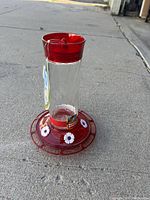 Single red hummingbird feeder standing outdoors on concrete, showing clear glass reservoir, red plastic base and top, and four white flower feeding ports.