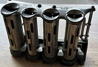Metal coin dispenser showing four vertical cylindrical compartments with slotted windows and a handle.