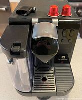 Front and top view of the black Nespresso coffee machine including two red coffee capsules on top and a small black accessory placed on the machine.