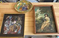 Three framed items on a table: wooden framed portrait, gold framed abstract artwork, and metal oval decorative plate.