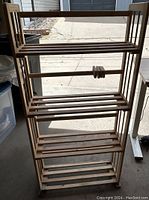 Wooden bookshelf shown from a slight side angle revealing the four wooden slatted shelves and vertical supports, placed on a concrete floor near other household items.