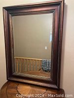 Rectangular mirror with wide dark brown wood frame reflecting interior staircase.