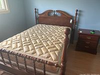 Full view of bed showing wood veneer headboard and footboard with turned posts, beige quilted mattress, box spring, and metal frame, beside a wooden nightstand.