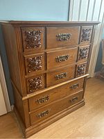 Frontal angled view of wood veneer dresser showing five drawers with decorative carved accents on top three drawers and metal handles.