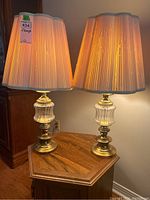 Pair of table lamps illuminated, showing the glowing fabric shades and brass and glass bases on a wooden table.