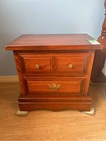 Front view of a medium brown wood veneer night stand showing two small top drawers with round metal pulls and one larger bottom drawer with a decorative metal pull, placed on a wood floor.