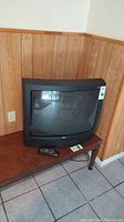 Front view of a black 27 inch vintage RCA CRT TV sitting on a wooden table against a wood paneled wall. Remote placed beside TV.