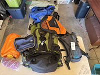 Multiple backpacks and bags arranged on table and floor in garage space including olive green, orange, blue, black, and grey colored items.