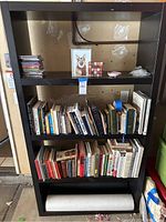 Full front view of black wooden bookshelf with three shelves containing books, CDs, and decorative items.