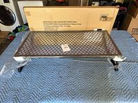 Folded campfire grill assembled on a blue padded surface showing mesh metal cooking surface and foldable legs.