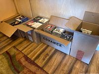 Multiple cardboard boxes on floor containing CDs, DVDs, and VHS tapes, showing variety and quantity