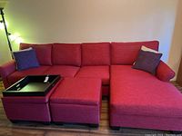 Front-facing photo of the full large red couch with chaise lounge, two storage cubes, a black tray on one cube, and two purple pillows at each armrest. Floor lamp visible to left.