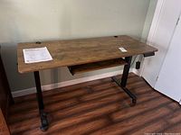 Front view of the wood desk supported by a black metal adjustable frame base, showing the floor and wall behind.