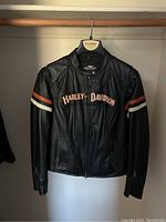Front view of black Harley-Davidson leather jacket hanging on wooden hanger, displaying front zipper, two zippered pockets, and orange and white stripes on sleeves with Harley-Davidson text on chest.