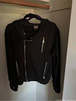 Front view of black Harley-Davidson women's zip-up sweater hung on a hanger, featuring two front pockets with zippers and a chest zipper pocket, zipped partially up, on a wooden hanger in a closet.