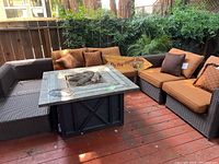 Wide view showing five-piece wicker sectional seating with cushions, gas fire pit table in center, decorative pillows and palm plant behind