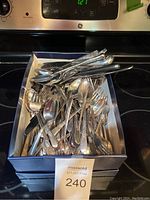 Large box filled with mixed silver plate and stainless steel flatware on a stove.