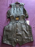Two black leather vests laid flat on a surface showing front view, one with side laces, the other with snap buttons and front pockets