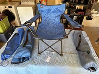 Photo showing one blue folding lounge chair with two matching blue carrying bags on a padded surface in a garage setting.