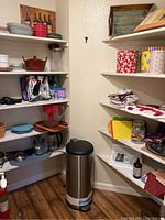 Photo of pantry corner shelving showing various kitchen plates, bowls, and cups along with a stainless steel trash can on floor.