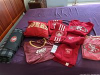 San Francisco 49ers branded apparel and accessories displayed on a purple surface including shirts, sweatshirts, scarves, and blanket.