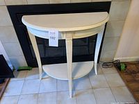 Front view of small half-round white side or entry table with lower shelf placed against fireplace on tile floor.
