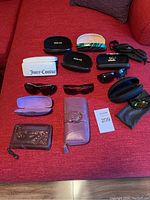 Photo of sunglasses, sunglasses cases, purple wallet, and brown zippered wallet spread on a red surface.