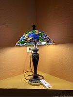 Full view of the Tiffany style lamp lit, showing the stained glass shade in blue, green, cream, purple, and pink colors with floral design and the dark metal base on a yellow surface.
