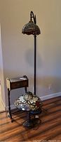 Photo showing Tiffany style floor lamp with hanging stained glass shade, Tiffany style table lamp with curved stained glass shade, and small wooden side table with drawer and shelf on hardwood floor against white wall.