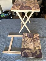 Photo of two wooden TV trays with sea life design on top, one standing upright and one folded, showing foldable crossed legs and surface decoration.