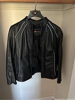 Front view of black Wilsons Leather jacket with white piping detail and zipper open showing removable liner attached inside.