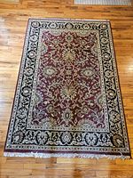 Full view of the rectangular burgundy area rug with black and gold floral border and white fringe on wood floor.