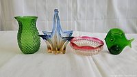 Four colorful handmade glass items: a green vase, a blue-amber star basket, a pink-rimmed trinket dish, and a green fish figure, all arranged on white cloth.