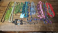 Top view of all 15 colorful beaded necklaces arranged across wooden surface showing variety in colors, lengths, and bead styles.