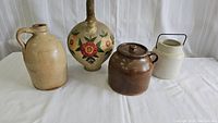 Four pieces of crockery on white cloth background showing jug with handle, floral painted jug, brown lidded jar, cream jar with metal handle.