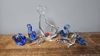 Seven handmade blown glass animal figurines on a wooden surface against a light gray wall showing cat, rabbit, seal, dolphin, squirrel, and bird in clear and blue colors.