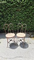 Pair of metal ice cream parlor chairs shown from the front with foliage background, showing rust and wear.