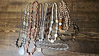 Lot of 12 natural tone necklaces laid out on a wooden surface, showing the variety of bead types, materials, and colors including large pendants and carved beads.