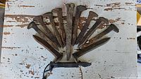 12 iron railroad spikes arranged in a fan shape on a white chipped paint surface. One spike is modified into a bottle opener with a lanyard attached.