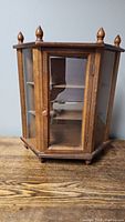 Closed view of small wooden curio shelf with hexagonal shape, wooden finials on top, and plexiglass sides and door.