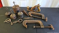 Photo showing the full lot on black surface including scale weights stacked with hooks, wooden hand plane, three large cast iron C-clamps, and various metal clamps and forged objects.