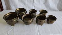 Seven vintage brass planters on feet displayed upright, showing variety in size and presence of handles on four.