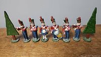 Front view of six painted toy soldiers and two painted tin trees arranged in a line, showing detailed uniforms and facial features.