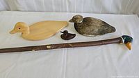 Four wooden duck-themed items laid flat: walking stick with duck head handle, large carved duck decoy, small dark brown carved duck, and duck-shaped charcuterie board