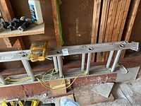 Aluminum extension ladder laying horizontally on garage floor, showing bumpers and eyelets attached with yellow rope.