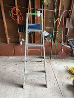 Front view of 5 ft aluminum step ladder with tool tray and orange extension cord hanging on wall in background.