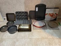 Image showing assortment of metal bakeware including muffin pans, cookie sheets, cake pans, cooling racks, and a glass casserole dish with lid, plus a boxed baking stone