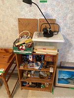 Photo showing wooden bookshelf filled with office supplies, desk lamp on top, cork boards behind, and additional items on shelves and floor.