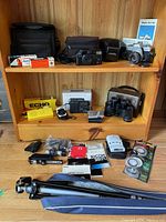 Photo of various items on wooden shelves including cameras with cases, binoculars, manuals, and accessories.