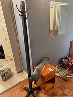 Wooden coat rack with multiple hooks on top and cross base; pine side table with drawer and rolled decorative mat on top; parquet flooring and adjacent wall